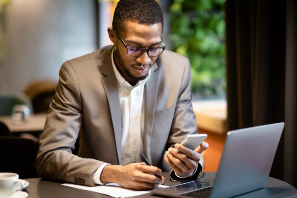 Black business guy taking notes in notebook holding phone