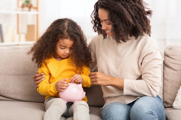 Little Girl Putting Money To Piggybank Sitting With Mother Indoor